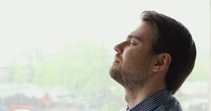 Close up side head shot view tranquil young man standing near window, breathing fresh air, dreaming or visualizing good future, copy space for motivating text, anxiety relief, no stress concept.