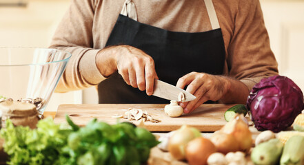 Crop unrecognizable bearded male in apron slicing champignons on wooden cutting board while preparing healthy dish with fresh ingredients in home kitchen.