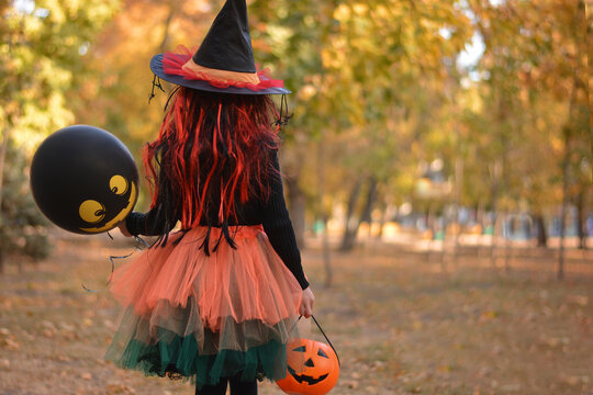 Little Girl In Witch Costume With A Balloon And A Pumpkin Bucket Playing In Autumn Park. Child Having Fun At Halloween Trick Or Treat. Kids Trick Or Treating.