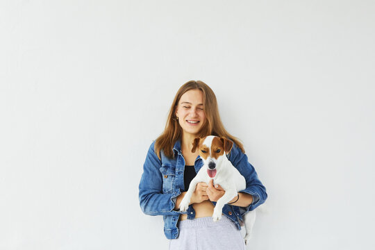 Young cheerful caucasian woman in a denim jacket is holding her pet, jack russell terrier, and is smiling with her eyes closed. Dog is looking into the camera with his tongue out. On white background.