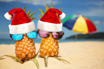 Couple of attractive pineapples in stylish mirrored sunglasses on sand against turquoise sea and beach umbrella. Wearing christmas hat. Christmas and new year vacation concept on tropical beach.