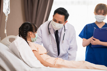 Fototapeta premium Asian male doctor consoling and talking with recovering girl patient during medical visit at hospital ward with female nurse standing in background. Medical workers visiting a girl patient.