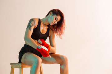 A young boxer girl with red bandages on her hands poses on a chair in a photo Studio.