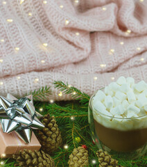 Christmas ornament. Gift box with ribbon, cup of cocoa with marshmallows, pine cones. Christmas tree branches. Light background with garland lights.