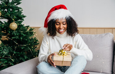 Portrait of smiling african American young woman in santa hat lying under decorated Christmas fir tree with wrapped gifts, happy biracial female congratulate greeting with new year on winter holidays