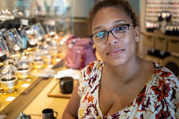 portrait of an African american black woman with glasses is sitting down in coffee cafe happily. she is wearing a colorful dress and waiting on her drink.