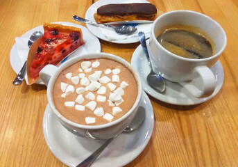 two cups of coffee on a wooden table: one cup of coffee with marshmallows, the second cup of coffee, two plates with a slice of berry pie and eclair in chocolate