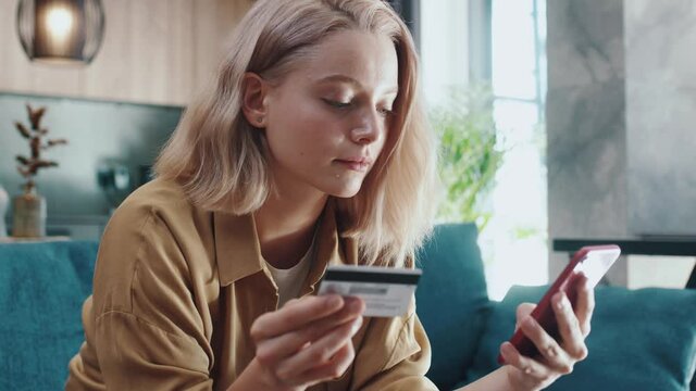 Attractive casual blonde woman using smartphone for distant transactions entering credit card number paying online shopping from home. E-commerce. Finance concept.