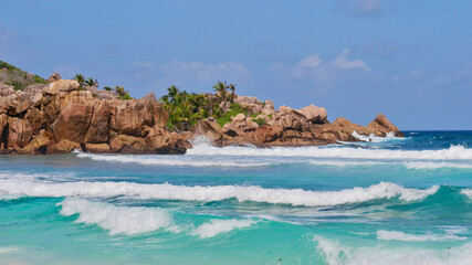 Fototapeta na wymiar Characteristic beautiful granite rock formations with coconut trees in between and breaking waves in turquoise water on beach Petite Anse in the south of La Digue island, Seychelles.