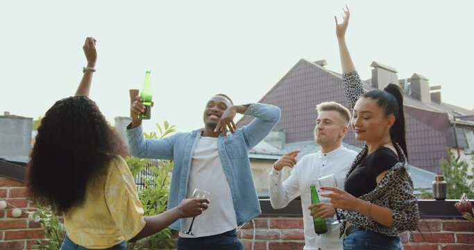 Portrait of good-natured smiling lucky 25s multiethnic friends which gethering together to celebrate summer party on the balcony with dances and beer and wine