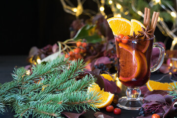Mulled wine with orange slices and spices on black wooden table with christmas lights and spruce branches in background. Selective focus, copyspace