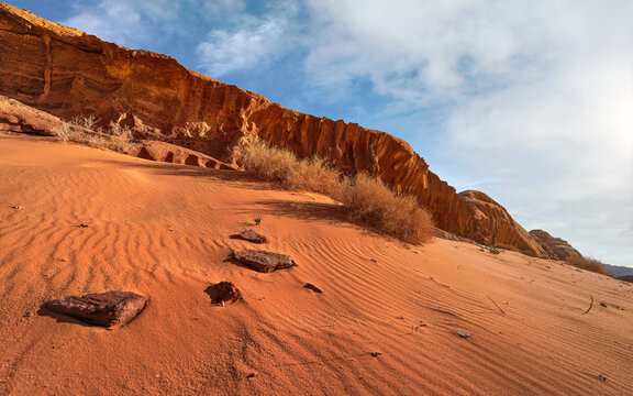 Rock formations in Wadi Rum desert, bright sun shines on red sand, rocks, some dry bushes, blue sky above