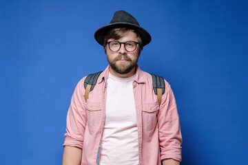 Caucasian bearded, shaggy man in hat, glasses and casual clothes looks calmly into the camera. Blue...