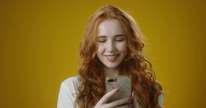 Cute authentic redhead caucasian girl using her smartphone, scrolling social media and positively smiling, shot on yellow background 4k footage