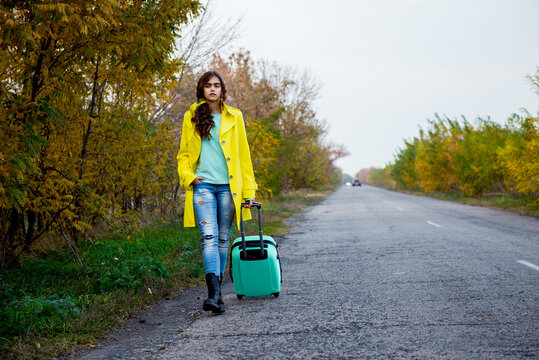 girl with a suitcase is walking along a road. The girl with wavy hair returns home. Young woman with a suitcase. Beautiful girl in a yellow raincoat in the fall. Hitchhiking on the road with luggage