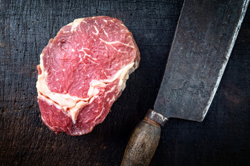 Raw dry aged angus rib eye beef steak offered as top view with a rustic old kitchen cleaver on an old charred wooden board
