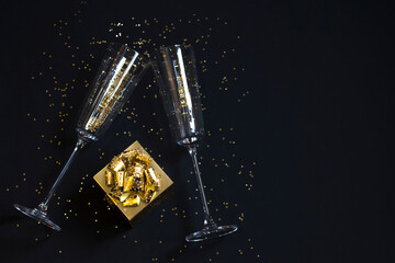 Gift box and two glasses, golden confetti on a festive black background. Festive Christmas, New Year concept. Copy space.