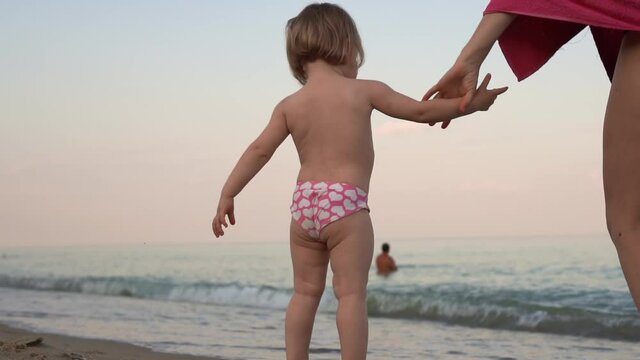 Small baby child girl standing at the beach looking at waves in the sea. Mother takes child by the hand. Family togetherness. Slow motion shot