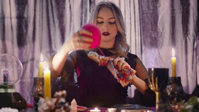 Dark magic voodoo witch performing ritual and casting spell piercing doll with needles sitting in dark room with burning candles.