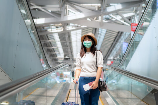 Asian traveler  woman with luggage wearing face mask looking outside terminal in airport standing on escalator go to gate for journey. New Normal travel concept.