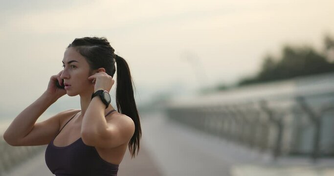 Sportswoman listening to music during training. Young female athlete looking at camera, listening to music in true wireless earphones during fitness workout in evening