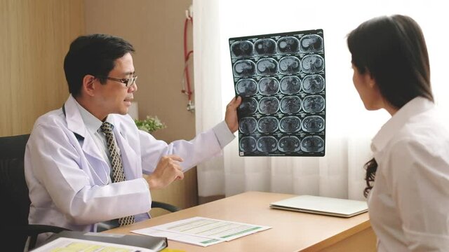 Asian doctor holding x-ray radiography film of the brain, explain the result to patient about migraine and brain cancer disease in hospital