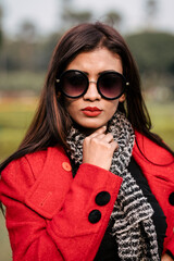 Outdoor fashion portrait of young beautiful fashionable female model wearing trendy long red winter coat, black suede ankle boots, scarf and sunglasses.