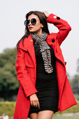 Outdoor fashion portrait of young beautiful fashionable female model wearing trendy long red winter coat, black suede ankle boots, scarf and sunglasses.