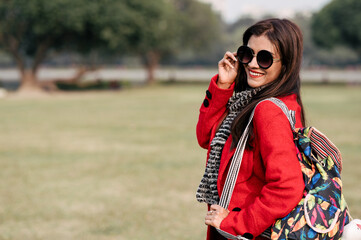 Education, high school and people concept - happy smiling young woman in glasses over green outdoor background. She is carrying her bag and wearing red dress for winter.
