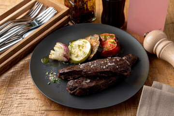 Roasted beef fillet, dark and yummy, with grilled vegetables as a side dish, knife, fork and pepper shaker aside on a wooden table