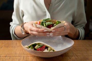 Woman is having lunch with pita, stuffed with salad and feta, wooden table, restaurant photo, no face is visible
