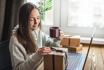 A happy girl in a sweater is sitting in front of a computer screen with a gift box in her hands. Concept of choosing gifts online and distance giving