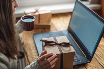A young woman is sitting in front of a laptop screen, using it and holding a gift box. Concept of online choosing and buying of gifts