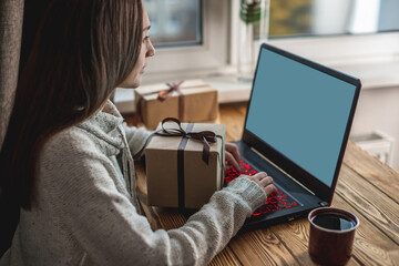 A young woman is sitting in front of a laptop screen, using it and holding a gift box. Concept of online choosing and buying of gifts