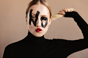 Woman with red lips and word no on face winks. Portrait of girl in black outfit holding her blond...