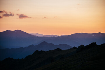 Warm gradient of dawn sky above layers of mountain and rock silhouettes. Vivid alpine landscape with dark rockies and orange sunrise sky. Minimalist highland scenery with silhouette of rocky mountains