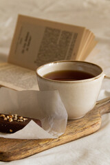 Cup with tea and a chocolate bar on a wooden board next to an open book on a white bed