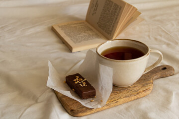 Cup with tea and a chocolate bar on a wooden board next to an open book on a white bed