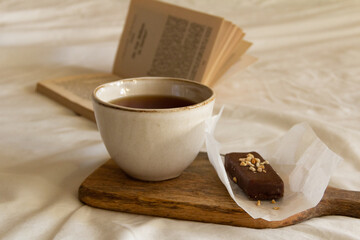 Cup with tea and a chocolate bar on a wooden board next to an open book on a white bed