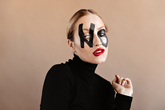 Woman Dressed In Black Sweater Coquettishly Posing On Beige Background. Snapshot Of Blonde With Word No On Her Face