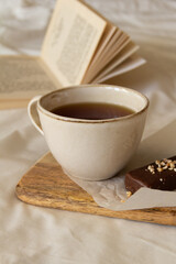 Cup with tea and a chocolate bar on a wooden board next to an open book on a white bed