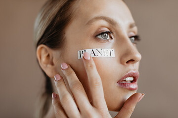 Lady without makeup touches her skin and looks into distance. Portrait of blonde girl with words planet on face
