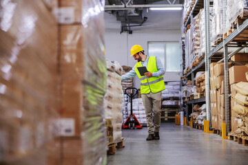 Full length of attractive bearded supervisor walking around warehouse and using tablet to check on...