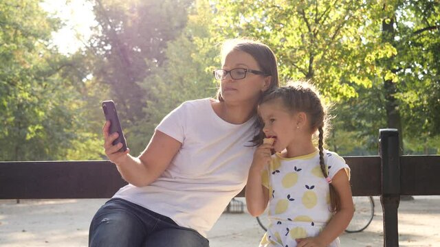 Woman mother makes phone photo selfie picture of her child daughter eat ice cream family together in city park sit on bench having fun
