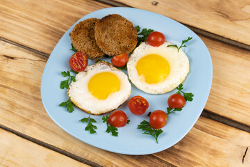 Fried eggs, bread croutons, cherry tomatoes, parsley on a blue plate. Wooden background.