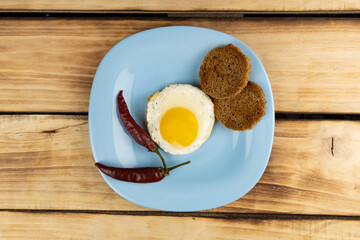 Top view of two chilli peppers fried egg and bread croutons on a blue plate. Wood background.