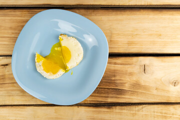 An overhead view of a fried egg cut in half, flowing yolk onto a blue plate. Wood background.