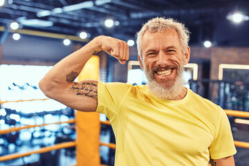 Portrait of a happy handsome mature man showing his biceps and smiling at camera while standing on...