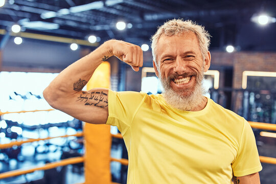 Portrait of a happy handsome mature man showing his biceps and smiling at camera while standing on the boxing ring at gym
