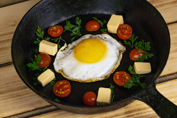 Top view of a fried egg, with slices of syrah, cherry tomatoes, herbs, on a black pan.